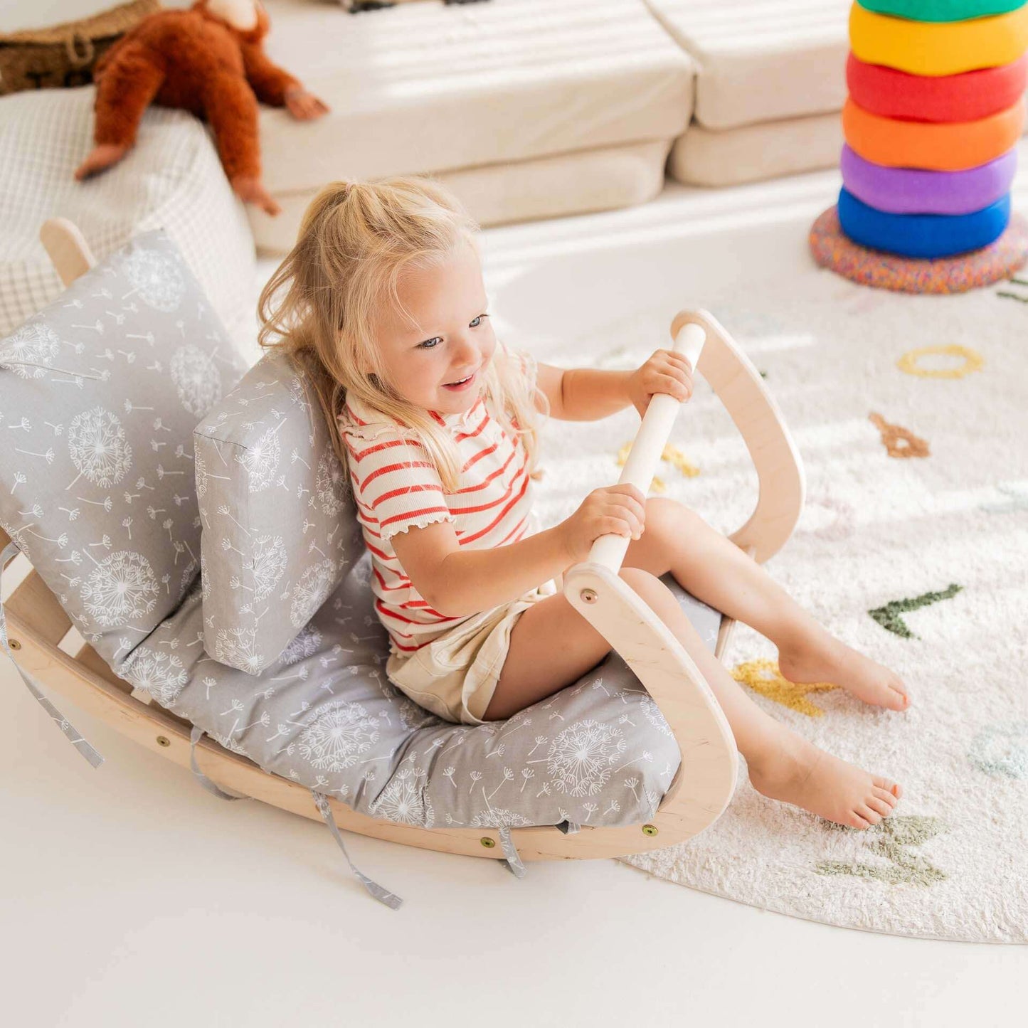 Kletterbogen Äffchen: Kind sitzt auf der Holz-Wippe und genießt das Schaukeln, umgeben von Kissen und einem bunten Teppich.