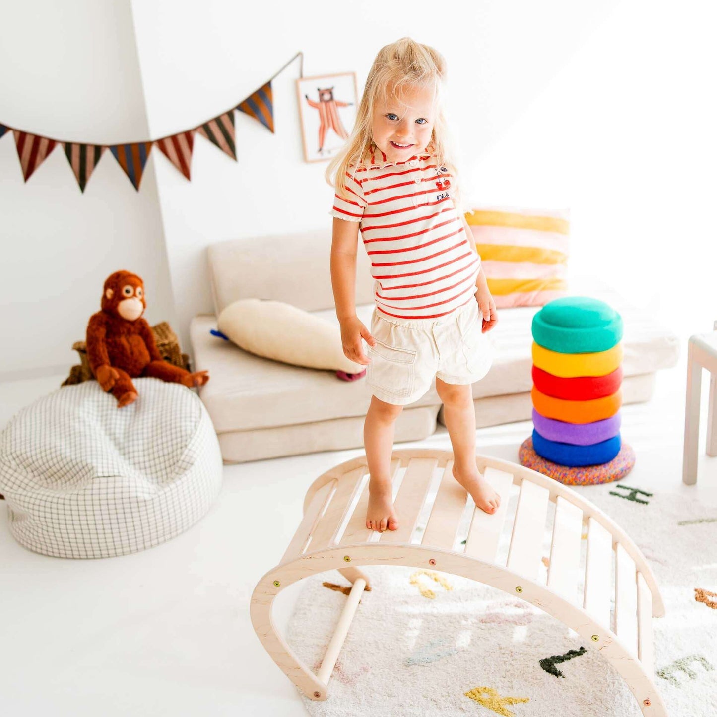 Ein kleines Mädchen steht auf einem Kletterbogen aus Holz in einem bunten Spielzimmer mit Spielzeug.