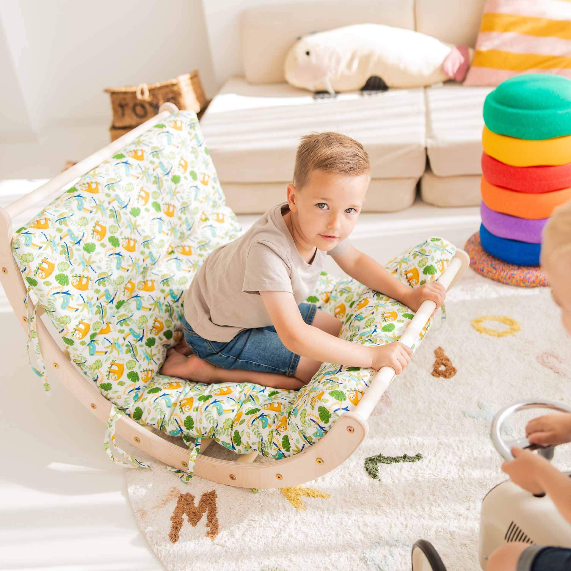 Ein Kind sitzt auf einem bunten Kletterbogen aus Holz mit Kissen, umgeben von Spielzeug und einer hellen, einladenden Umgebung.