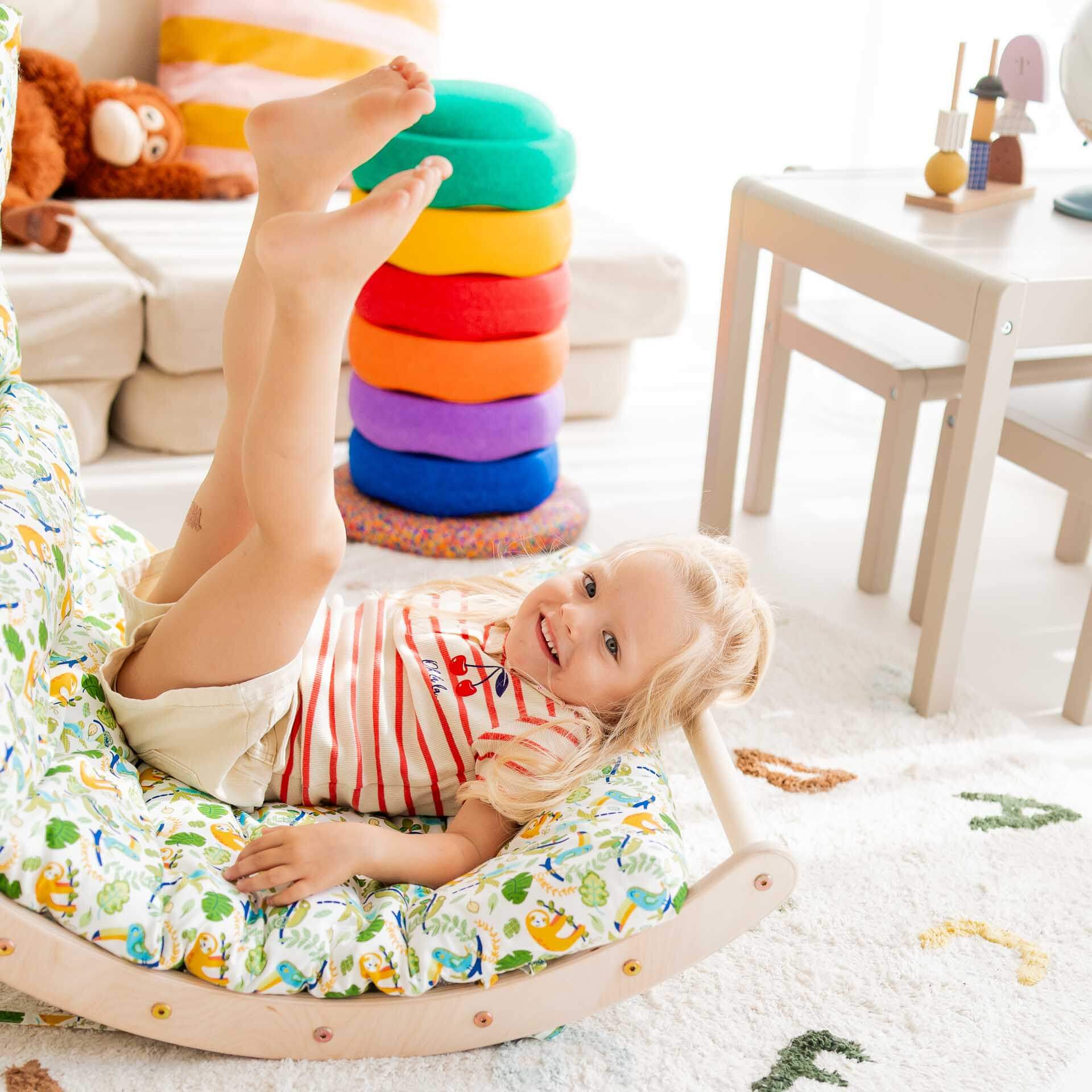 Fröhliches Kind spielt auf dem Kletterbogen aus Holz, umgeben von bunten Kissen und Spielzeug.